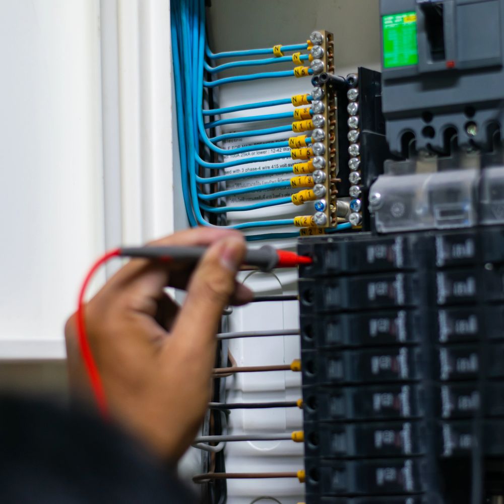 Hand using a multimeter probe on an electrical panel with labeled blue wires.