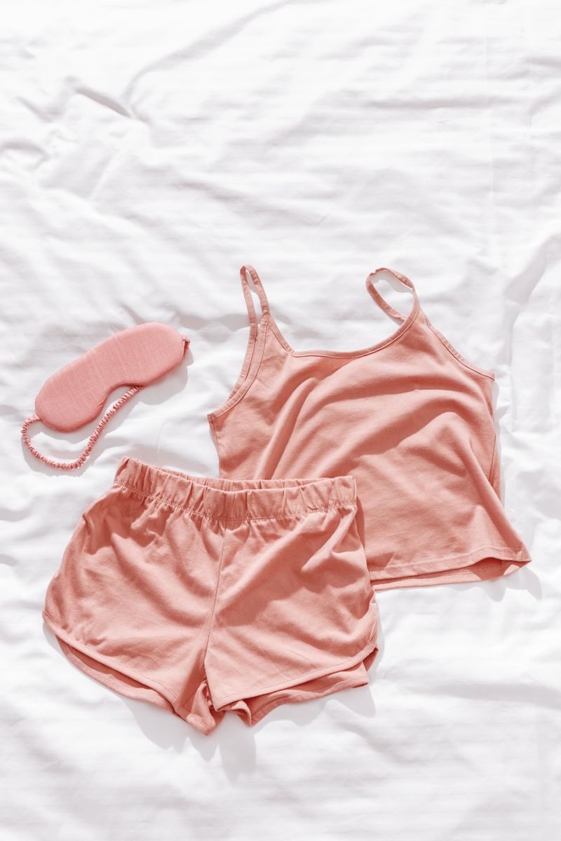 Pink female pajama and sleep eye mask on white cloth bed sheet. Top view comfort pyjamas for health sleeping. Romance mood, singlet and shorts for girl light pink trend color, flat lay with copy space