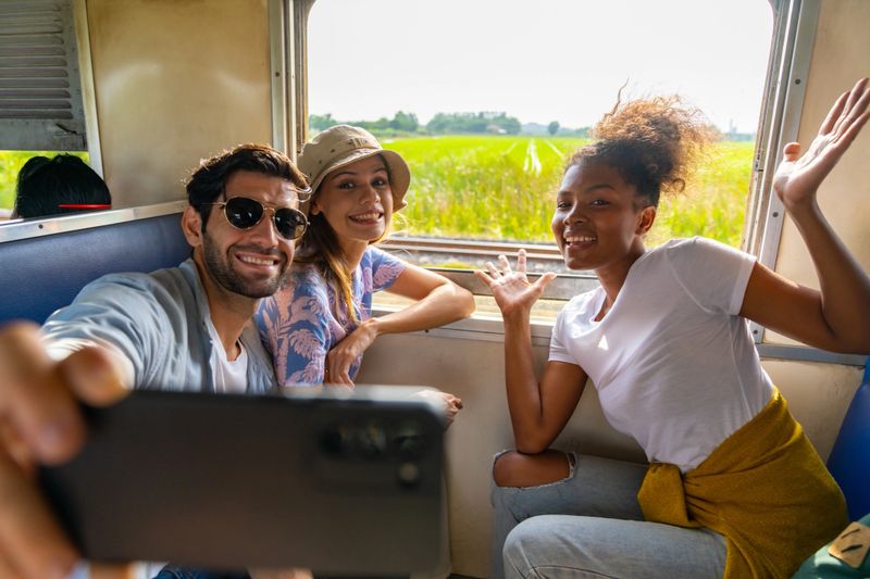 Happy Diversity people tourist travel in Thailand on local train. Man and woman friends using mobile phone taking selfie together during travel on railroad transportation on summer holiday vacations.