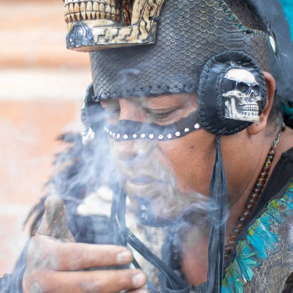 Indigenous man in traditional attire with skull face paint and smoke.