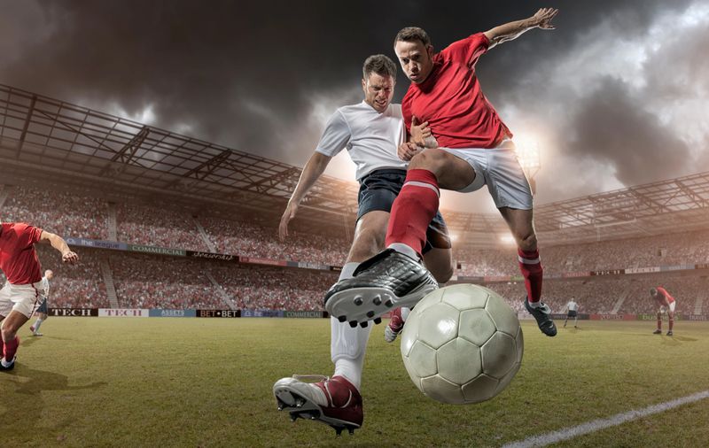 Mid-air action shot of professional footballers tackling on pitch during game in full floodlit stadium under stormy sky