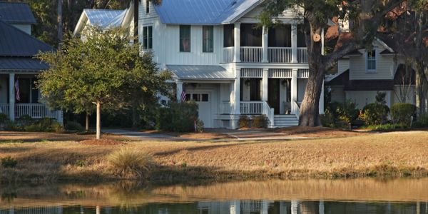 White two-story house with a porch by a pond and trees.