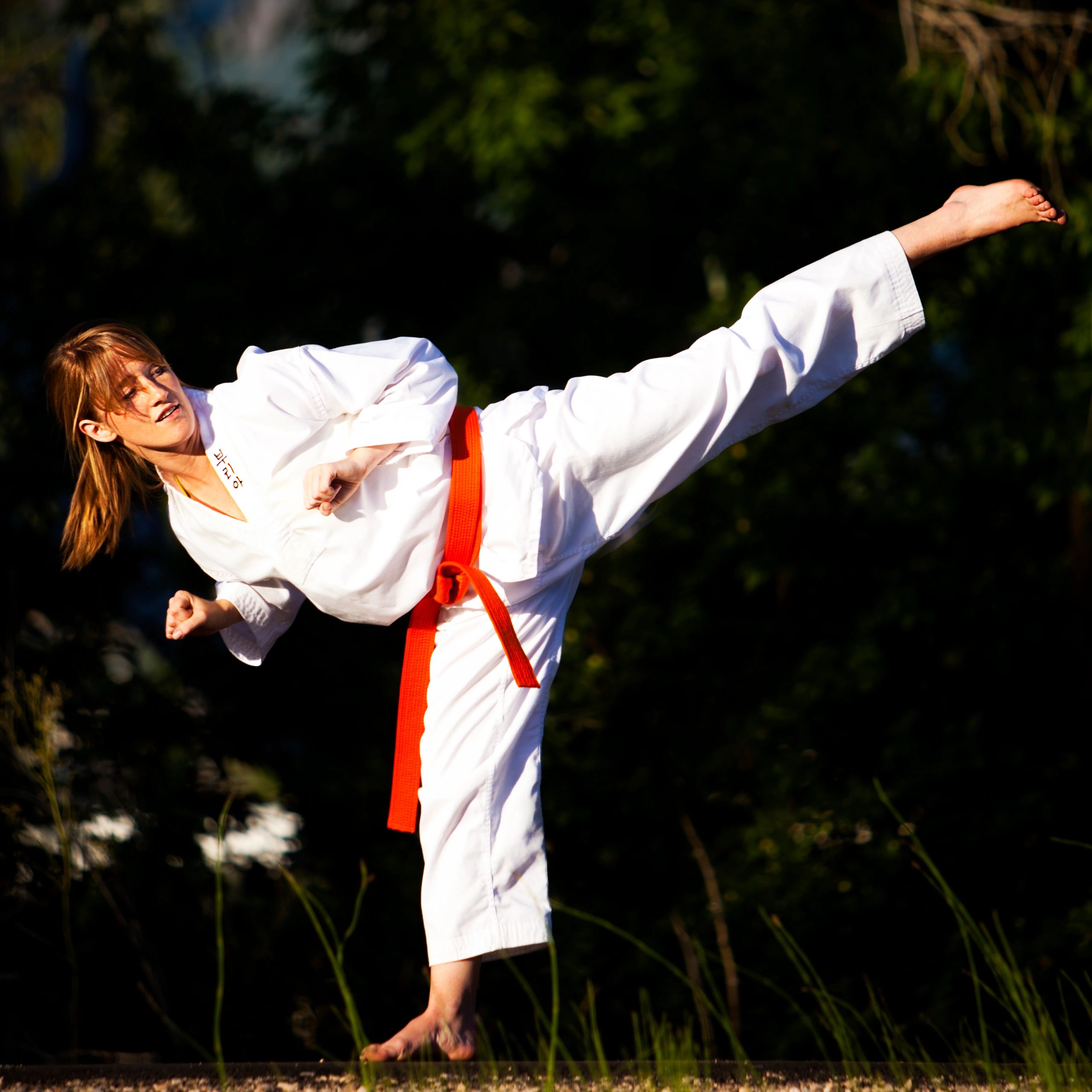 Woman in a white martial arts uniform performing a high kick outdoors.
