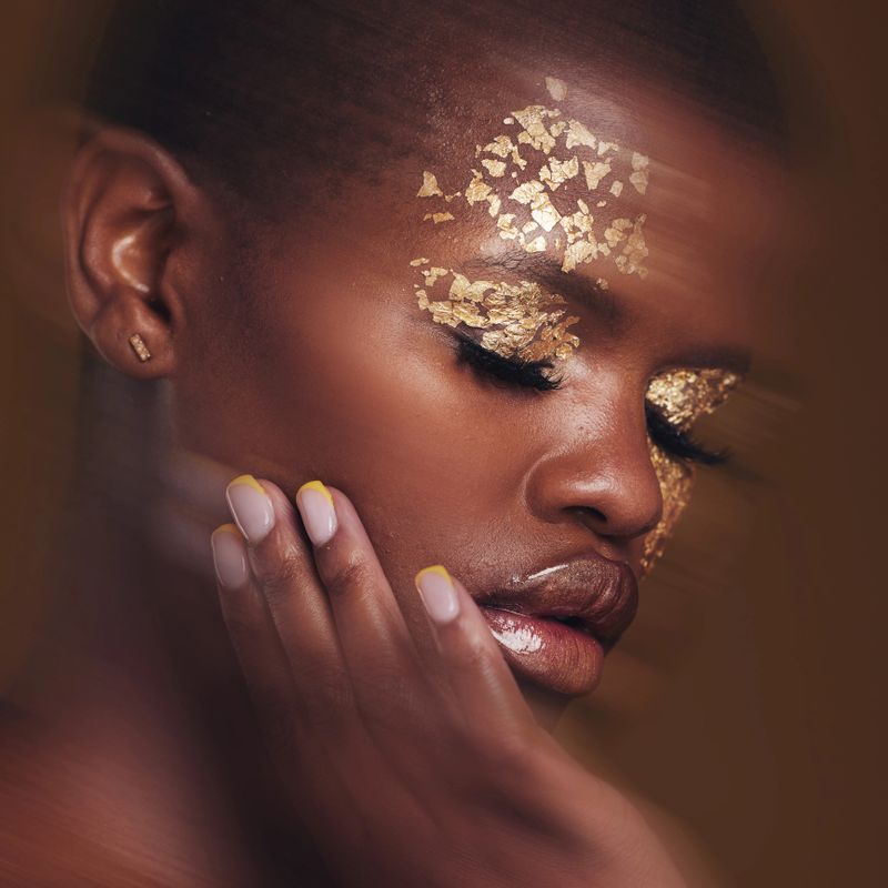 Gold beauty, blur and black woman with hand on face closeup with makeup and cosmetics in studio. Brown background, African female model and manicure with golden shine and glow from paint sparkle