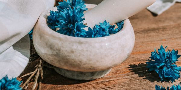 Hand dipping blue flowers into a stone bowl on a wooden surface.
