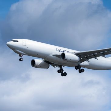 White cargo airplane flying with landing gear extended under cloudy sky.