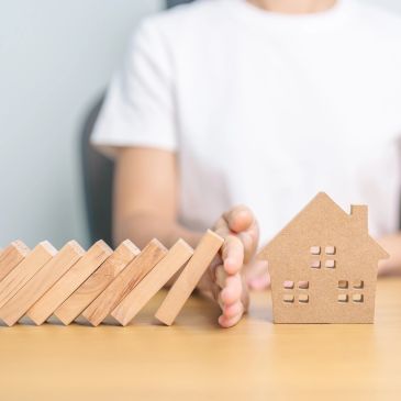 Hand stopping falling wooden blocks from hitting a cardboard house.