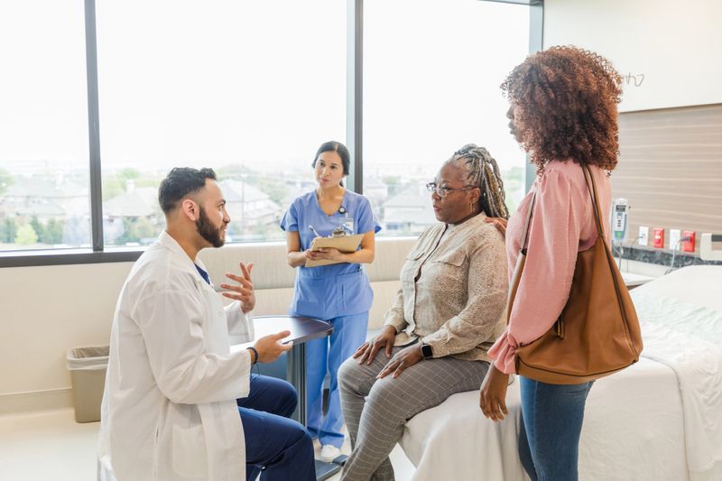 The young adult male doctor gestures as he talks to the mature adult female patient.  The patient's mid adult daughter and the mid adult female nurse pay close attention.