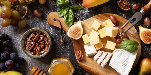 A wooden board with cheese cubes, figs, grapes, and pecans on a dark surface.