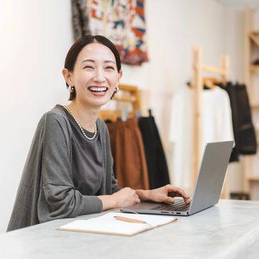 A joyful woman working on a laptop in a cozy workspace with clothes in the background.