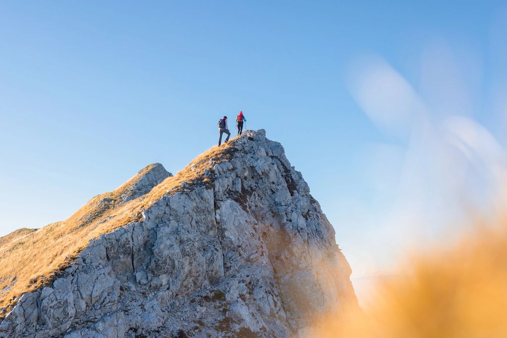 Two hikers reaching the peak of a rocky mountain under clear blue skies.