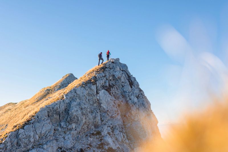 Spectacular view of two hikers walking along the mountain ridge route with the sun on the clear blue sky, aerial shot. Inspiration, nature, and mountaineering concepts.