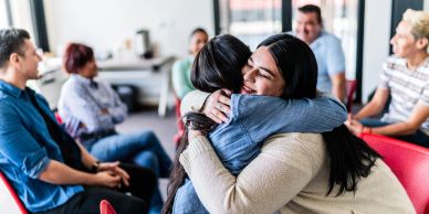 Two women hugging in a group setting with others watching.