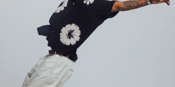 Man in tie-dye shirt and white pants leaning backward against a plain background.