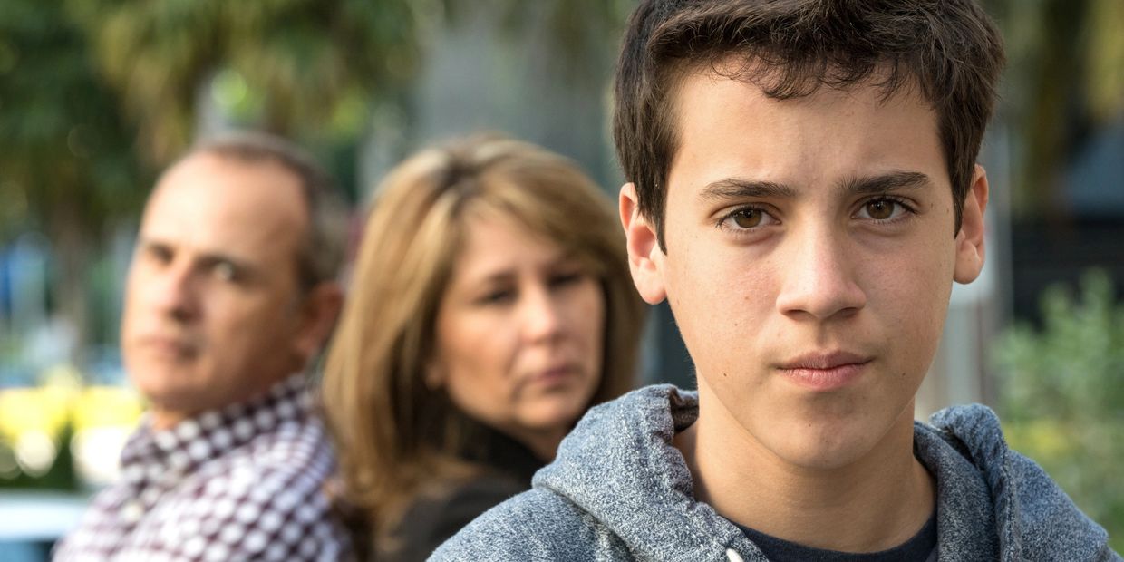 Teen boy looks serious with parents blurred in background.