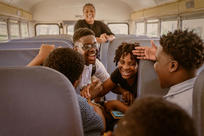 Yellow school drops students off at school. Students talk and interact with each other. Upon arrival they greet the principal of the school.