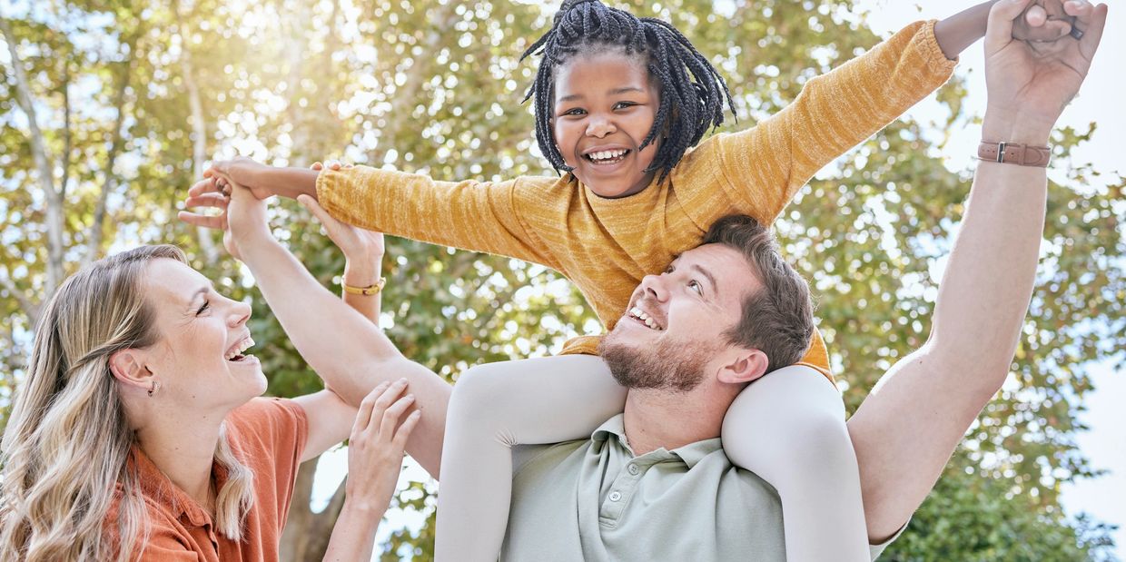 Happy family playing outdoors with a child on the father's shoulders, all smiling.