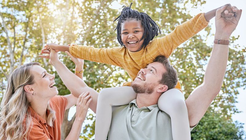 Family, adoption and piggyback in the park with a girl and foster parents having fun together in the park. Diversity, playful and freedom with a mother, father and daughter bonding outdoor in nature