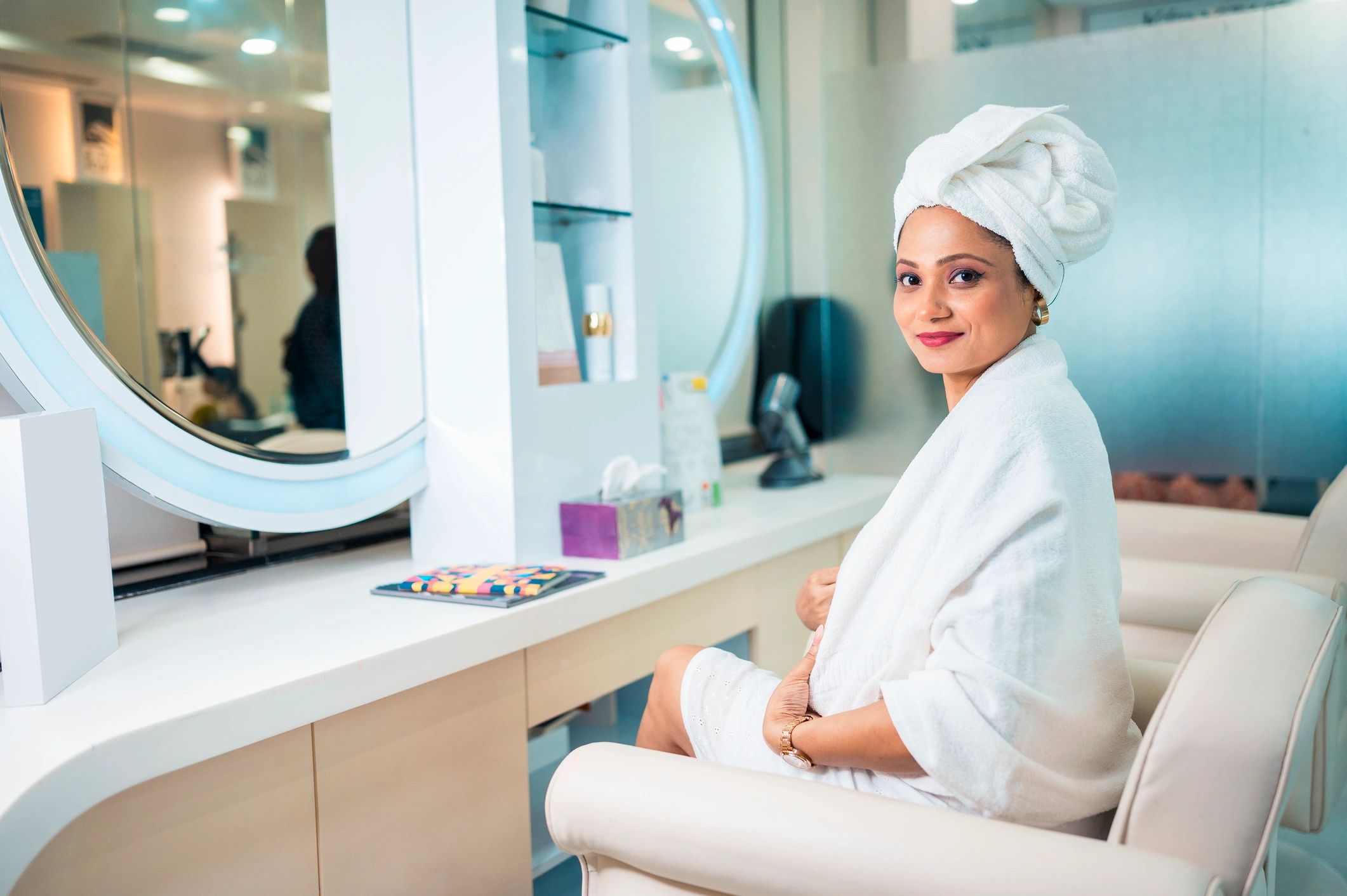 Woman in white bathrobe with towel on head smiling indoors.