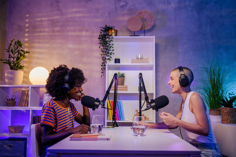Two happy multiracial young women are sitting in a podcast studio and live-streaming their discussion. Gen z girls are making a podcast. Two women wearing headphones and talking at the microphone.