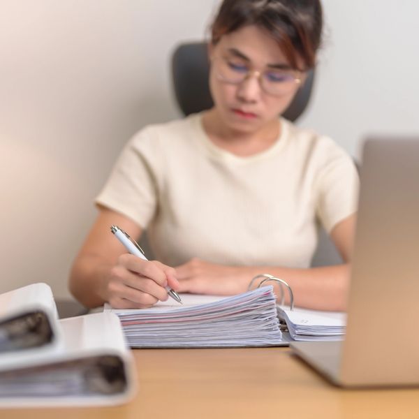Person writing notes beside a laptop with thick binders on the desk.