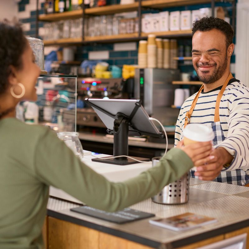Man With Down Syndrome Serving Female Customer At Coffee Shop Checkout