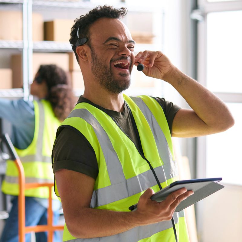 Man With Down Syndrome Working In Warehouse Wearing Headset Using Digital Tablet