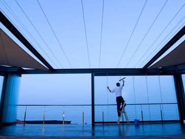 A man stands on a ladder cleaning large glass windows at dusk.