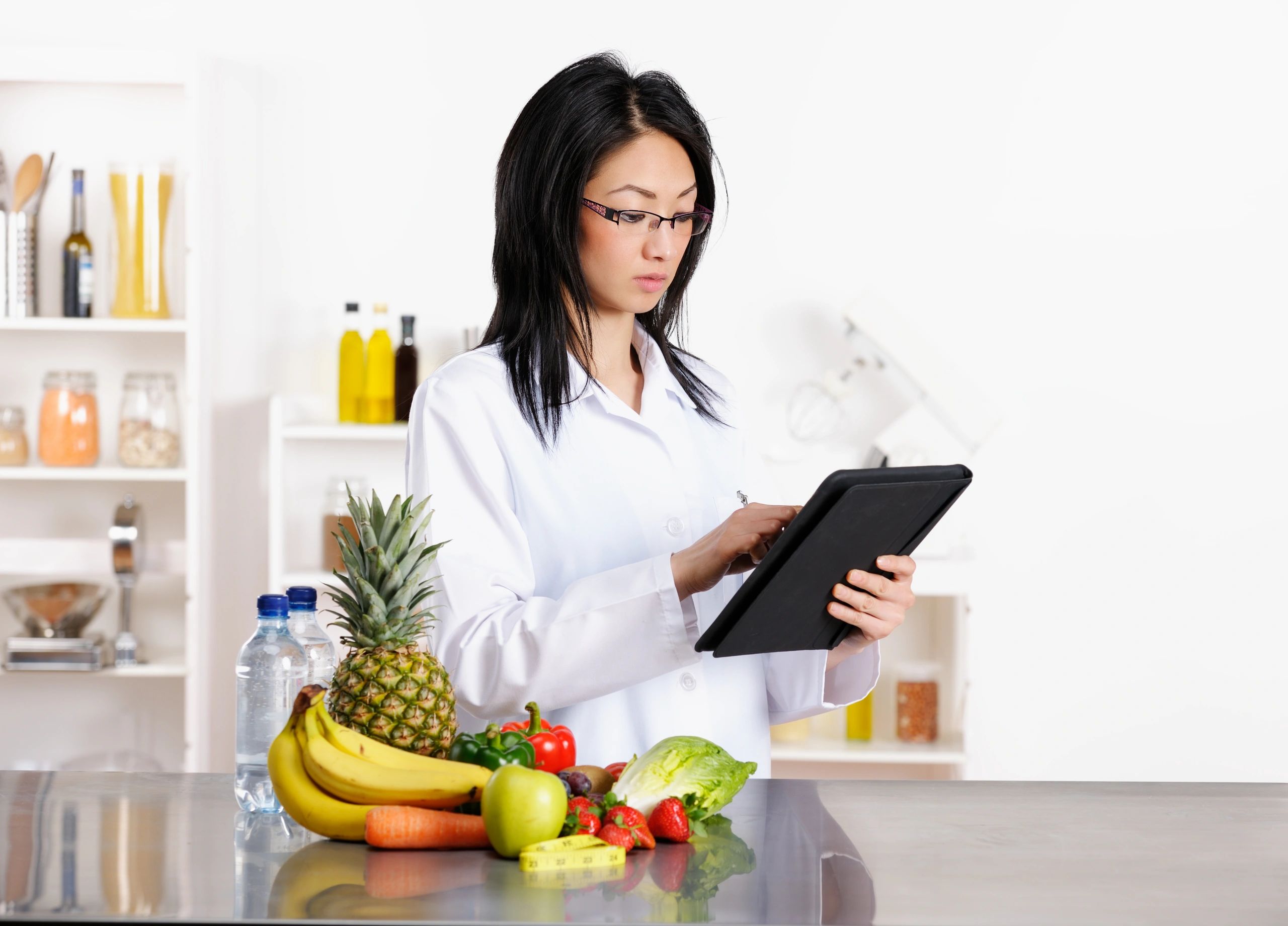 A female nutritionist in a lab coat checks a tablet with fresh fruits and vegetables on the counter.