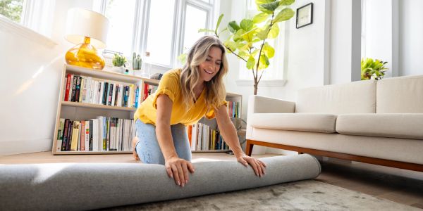 A woman happily rolling up a gray rug in a bright living room.