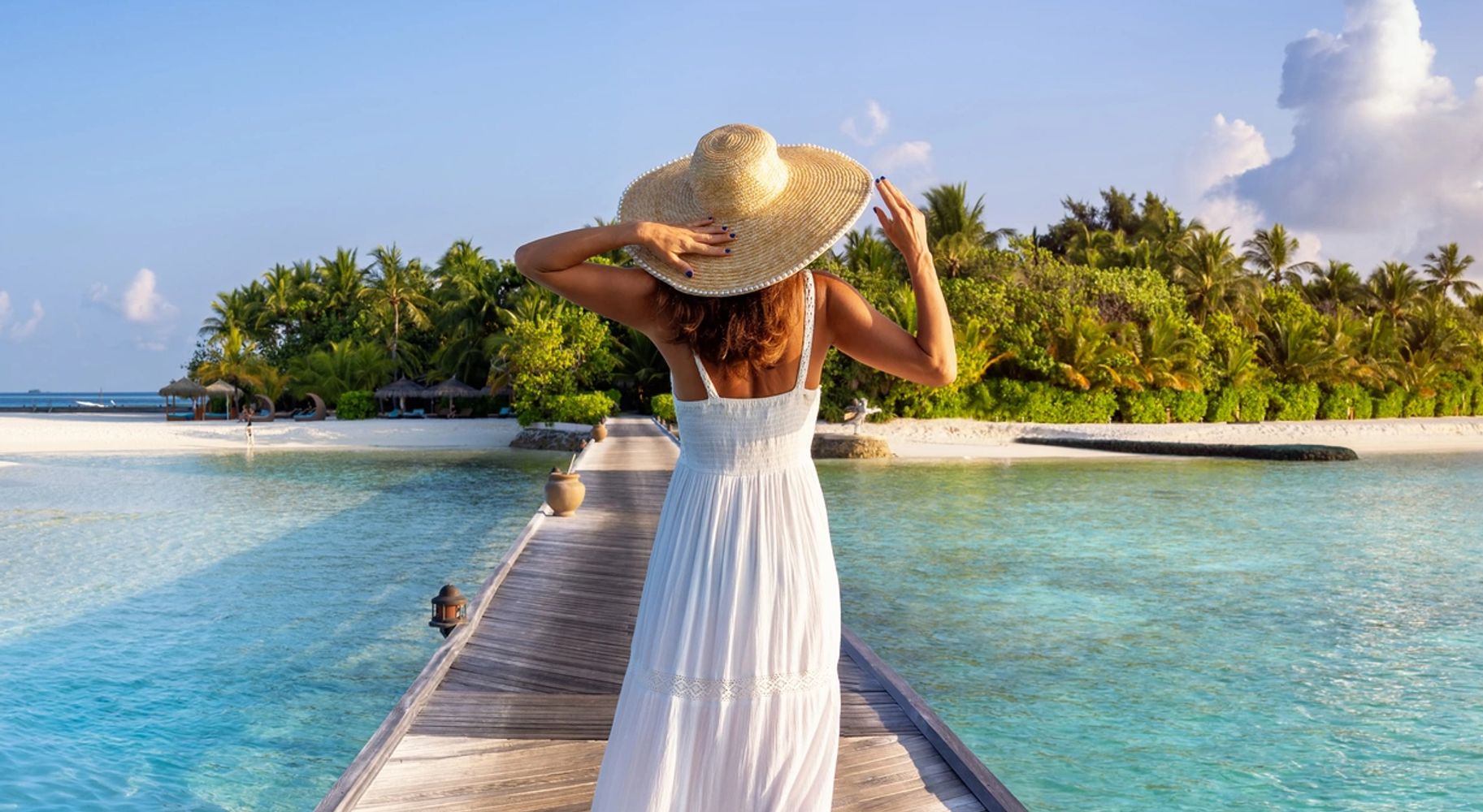 Woman in white dress and hat walking on a wooden pier towards a tropical island.