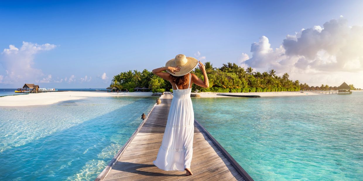 Woman in white dress and hat walking on a wooden pier towards a tropical island.