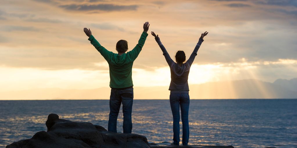 Two people raising arms by the ocean at sunset.