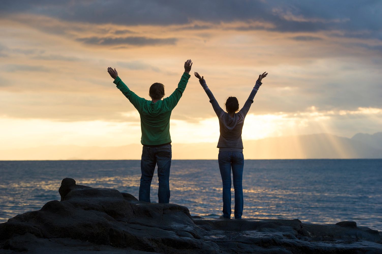 Couple standing on a coastal rock with their arms raised, looking towards the sun