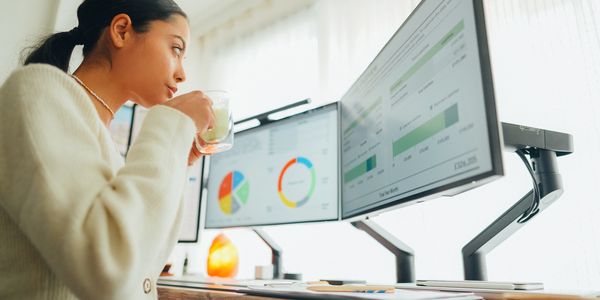 Woman drinking tea while analyzing charts on dual monitors at her desk.