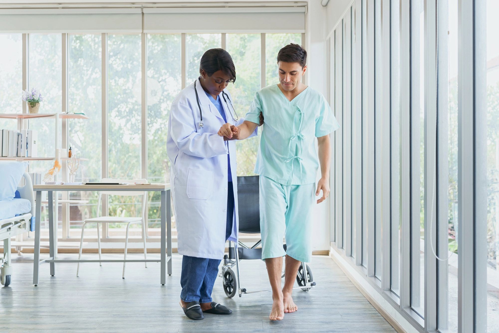 Doctor assists patient walking barefoot in a hospital hallway.