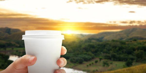 Hand holding a white coffee cup with a scenic sunset and green hills in the background.
