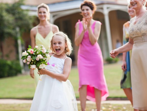 Excited flower girl holding a bouquet with bridesmaids clapping behind her.