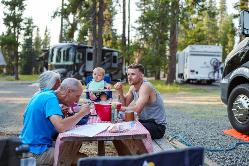 Active multi-racial senior couple sit with their multi-generation family at a picnic table in a campground and enjoy eating a meal outdoors while camping in the Pacific Northwest region of the United States.