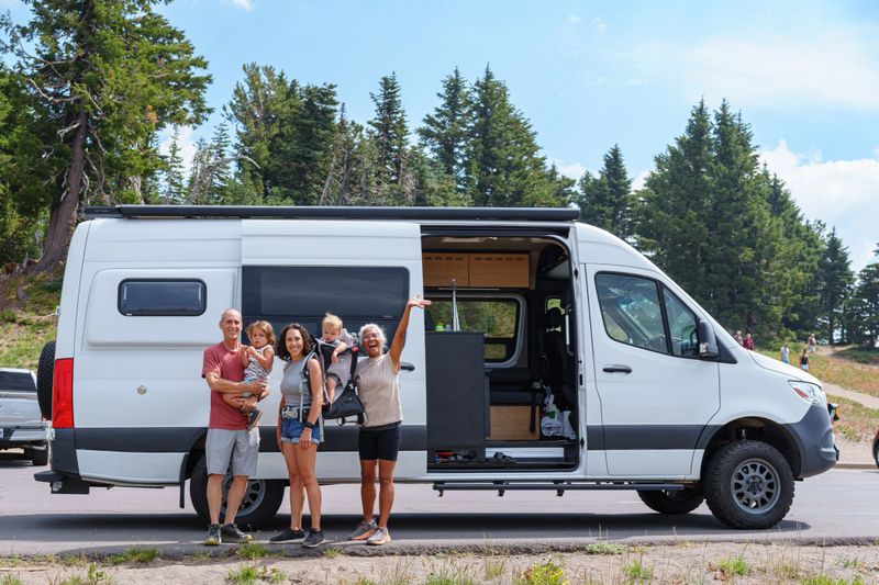 An active and loving multi-racial multigenerational family happily poses outside a camper van before going for a hike while on a summer camping road trip in Oregon.