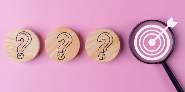 Magnifying glass highlights a target next to wooden blocks with question marks on pink background.