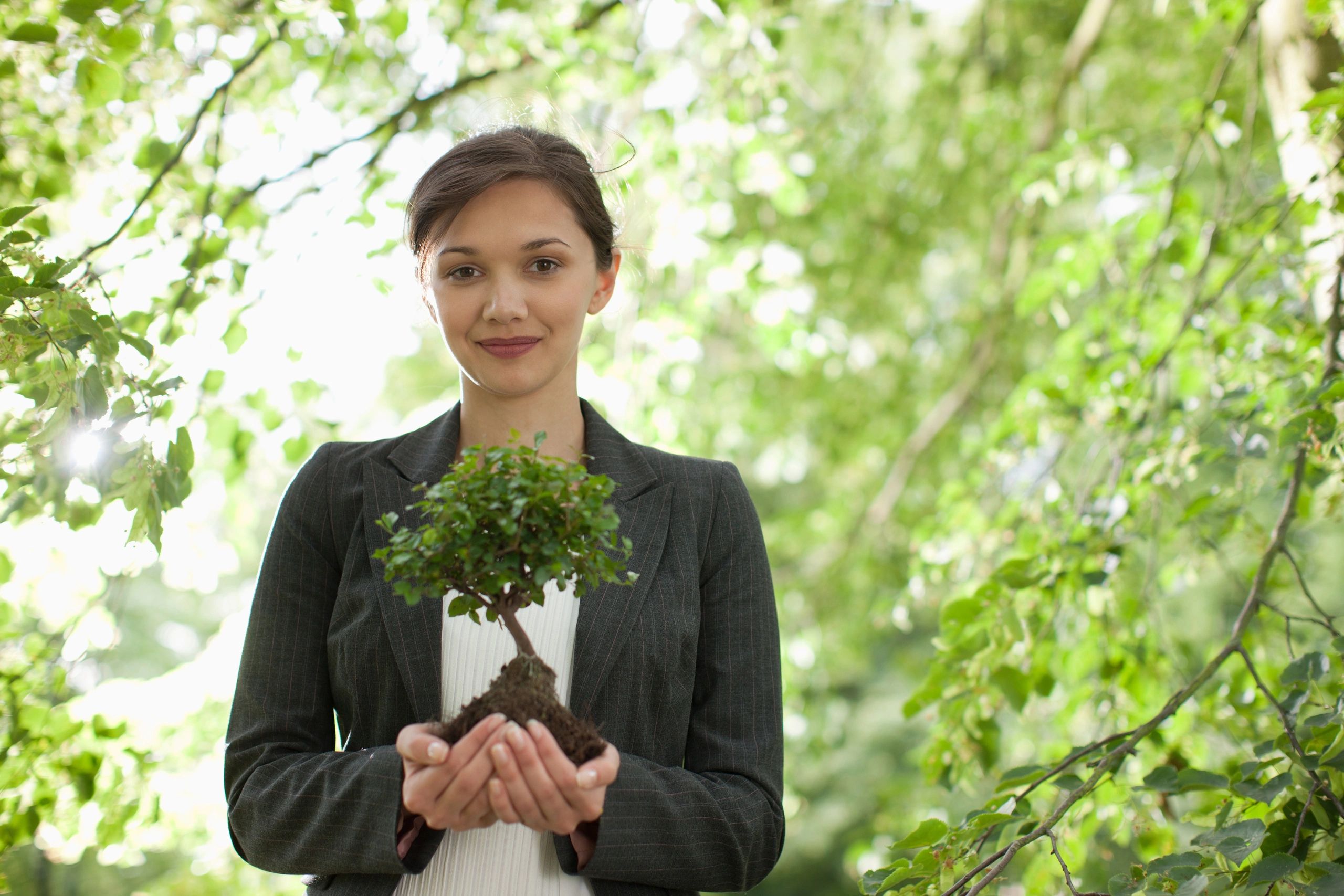 Woman holding a small tree with soil in a lush green environment.