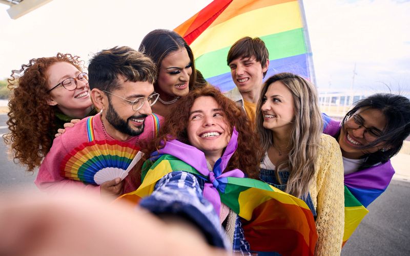 Fun selfie LGBT group young friends celebrating gay pride day together holding rainbow flag. Homosexual people community smiling and taking cheerful portrait outdoor. Generation z positive relations.