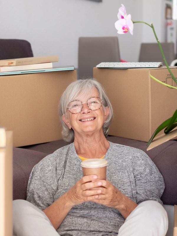 Elderly woman sitting with coffee cup among moving boxes.