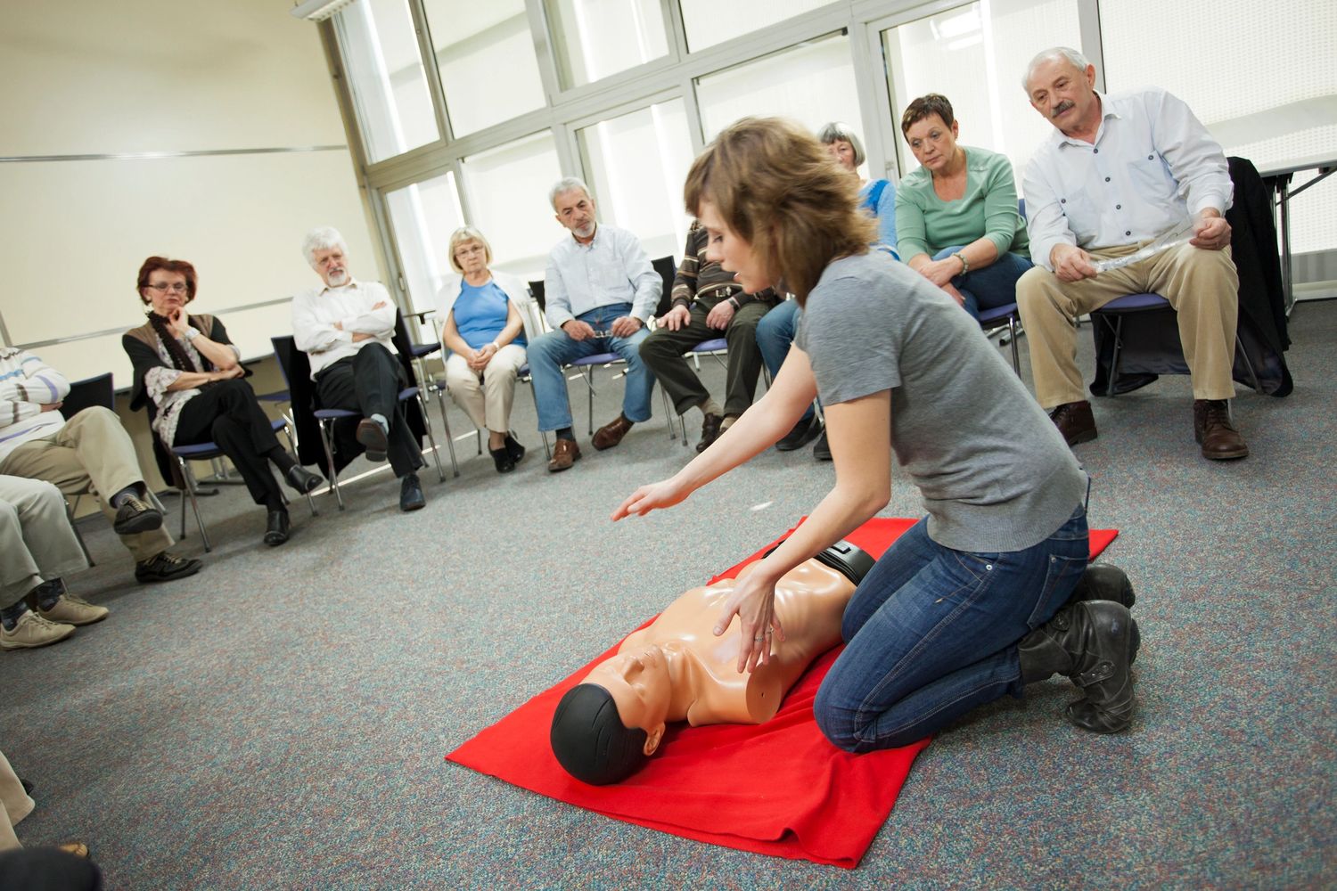 Woman demonstrating CPR on a mannequin to a seated group of adults.