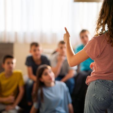 A girl speaking to a group of attentive teenagers indoors.