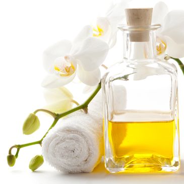A glass bottle of yellow oil, white orchid flowers, and a rolled towel on a white background.