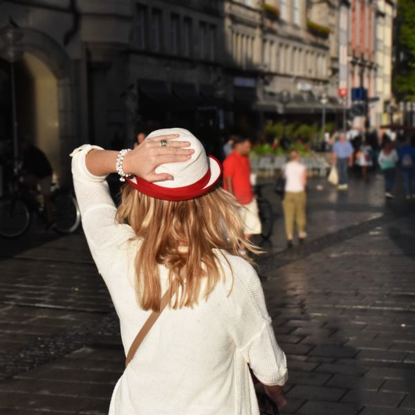 A woman adjusts her hat while walking in a sunny city street.