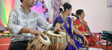 Musicians playing tabla and singing in traditional attire at a cultural event.