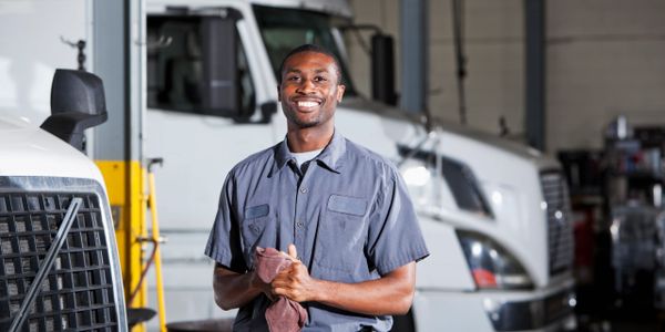 Smiling mechanic in uniform stands in front of large trucks in a garage.